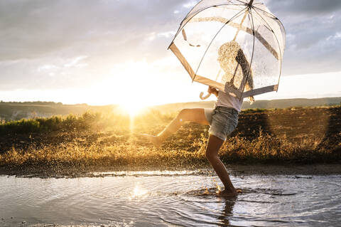 Mädchen mit transparentem Regenschirm spritzt Wasser auf Pfütze gegen Himmel bei Sonnenuntergang, lizenzfreies Stockfoto