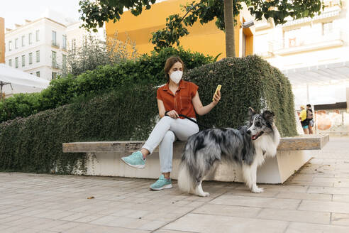 Young woman wearing protective mask sitting on bench in the city taking photo with smartphone - AGGF00085