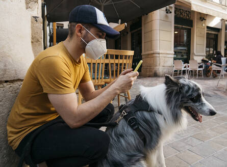Man wearing protective mask waiting with his dog in the city using smartphone - AGGF00088