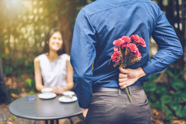 Rear View Of Boyfriend Hiding Flowers While Standing In Front Of Girlfriend In Garden - EYF07796