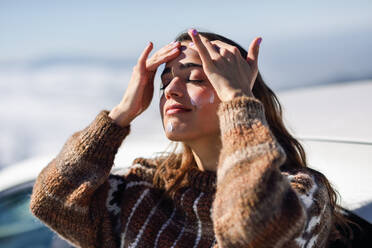 Junge Frau mit geschlossenen Augen trägt Feuchtigkeitscreme auf, während sie im Winter im Freien steht - EYF07823