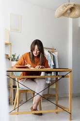 Female fashion designer working at home sitting at desk taking notes - ERRF04015