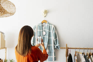 Weibliche Modedesignerin, die zu Hause arbeitet und mit ihrem Smartphone ein Foto von einem Kleidungsstück macht - ERRF04026
