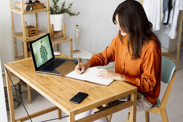 Female fashion designer working at home sitting at desk taking notes - ERRF04029