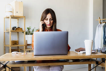 Female fashion designer sitting at desk in home office talking on the phone - ERRF04048