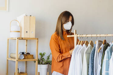 Female fashion designer working at home with clothes stand wearing protective face mask - ERRF04056