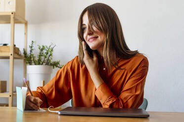 Female freelancer sitting at desk in home office talking on the phone and taking notes - ERRF04065