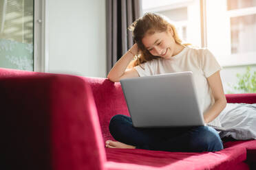 Lächelnde junge Frau, die einen Laptop benutzt, während sie zu Hause auf dem Sofa sitzt - EYF07861