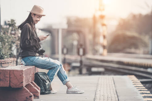 Lächelnde junge Frau, die ein Smart Phone benutzt, während sie auf einer Bank am Bahnsteig sitzt - EYF07866