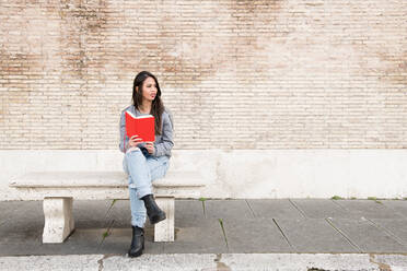 Junge Frau, die ein Buch liest, während sie gegen eine Backsteinmauer sitzt - EYF07925