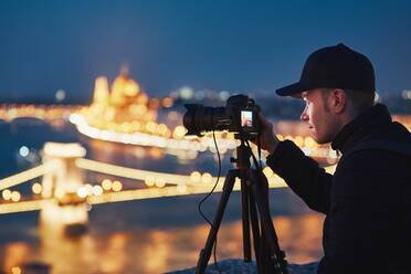 Mann fotografiert beleuchtete Kettenbrücke von der Kamera in der Stadt bei Nacht - EYF07931