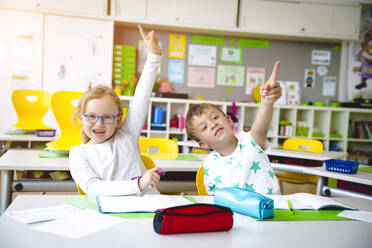 Junge und Mädchen mit erhobenen Händen am Tisch im Klassenzimmer - EYF07983