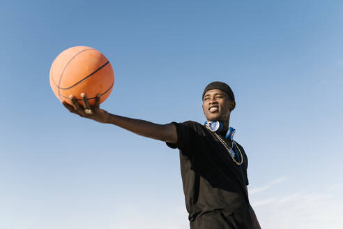 Junger Mann, der einen Basketball hält, während er an einem sonnigen Tag vor einem klaren blauen Himmel steht - EGAF00283