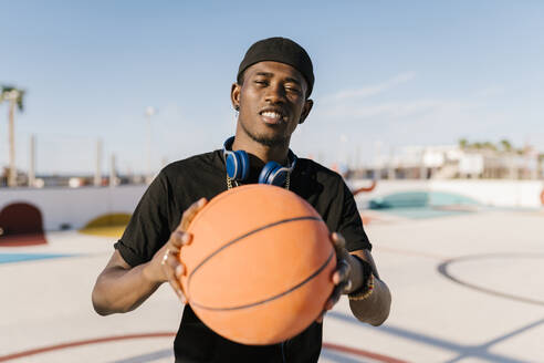 Junger Mann hält Basketball, während er an einem sonnigen Tag vor einem klaren Himmel steht - EGAF00284