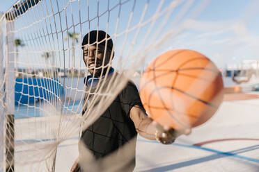 Junger Mann, der einen Basketball am Netz hält, während er an einem sonnigen Tag im Hof steht - EGAF00285