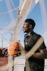 Young man holding basketball looking away while standing by net in court - EGAF00286