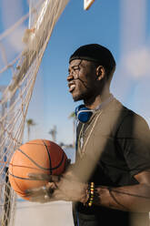 Man holding basketball looking away while standing by net in court during sunny day - EGAF00287