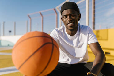 Smiling young man holding basketball while sitting in court during sunny day - EGAF00291