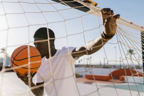 Junger Mann küsst Basketball, während er am Netz im Gericht steht, während eines sonnigen Tages - EGAF00293