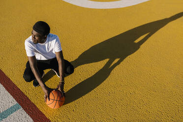 Thoughtful young man holding basketball crouching on sports court during sunny day - EGAF00294