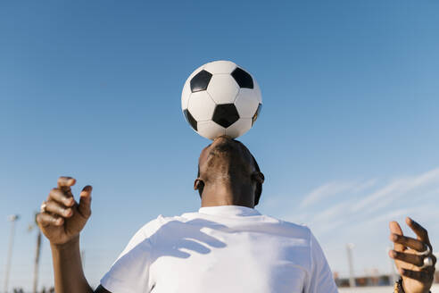 Nahaufnahme eines jungen Mannes, der einen Fußball auf dem Kopf gegen den blauen Himmel balanciert - EGAF00297