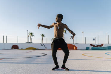 Shirtless young man dancing against clear sky in sports court - EGAF00300