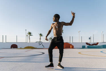 Shirtless young man dancing against clear sky in sports court during sunny day - EGAF00301