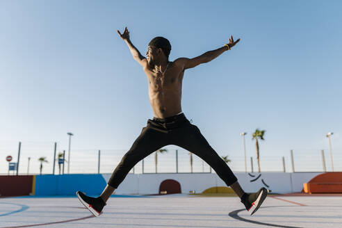 Shirtless young man with arms raised jumping against clear sky in sports court - EGAF00303