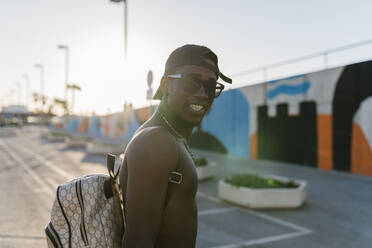 Shirtless young man wearing sunglasses standing on road during sunny day in city - EGAF00305