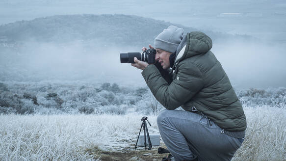 Seitenansicht eines Mannes, der im Winter auf einem Feld hockend fotografiert - EYF08079