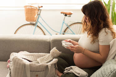 Schwangere Mid Adult Frau suchen Baby-Kleidung beim Sitzen auf dem Sofa zu Hause - EYF08108