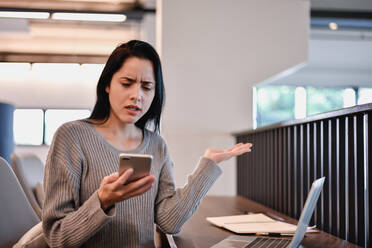 Frustrated Young Woman Using Phone While Sitting At Table - EYF08133