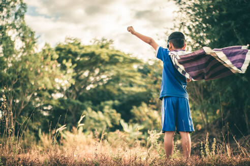 Rear View Of Boy Wearing Cape Standing On Grassy Field Against Trees - EYF08143