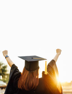 Rear View Of Woman Wearing Graduation Gown Standing With Arms Raised Against Sky - EYF08150