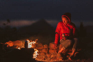 Female camper stares into fire while drinking beer on blm land of utah - CAVF85907