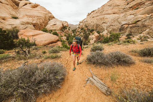 Frau stapft mit dem Rucksack durch Sand unter Sandsteinschluchten der Canyonlands - CAVF85962