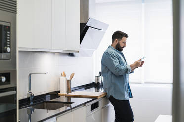 Mann kocht Crêpes in der Küche mit einem Mobiltelefon in einem Jeanshemd - CAVF85971