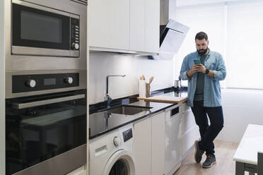 Man cooking crepes in the kitchen with a mobile phone in a denim shirt - CAVF85973