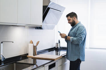 Man cooking crepes in the kitchen with a mobile phone in a denim shirt - CAVF85974