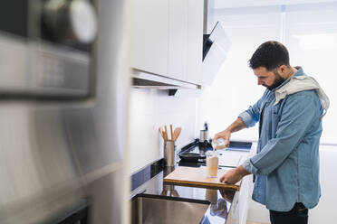 Ein Mann im Jeanshemd kocht in der Küche und schüttet Milch in einen Behälter - CAVF85986