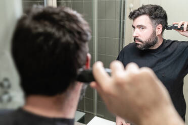 Selbsthaarschnitt: Ein Mann schneidet sich im Badezimmer die Haare mit einer Haarschneidemaschine - CAVF86001