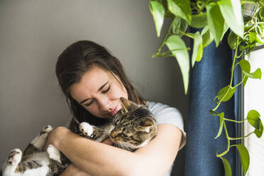 Woman indoors at home cuddling with her cat by the window - CAVF86014