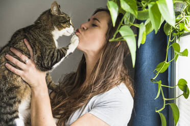 Woman indoors at home kissing her cat by the window - CAVF86015