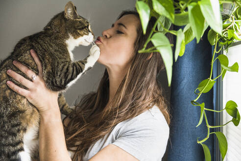 Frau zu Hause, die ihre Katze am Fenster küsst - CAVF86015