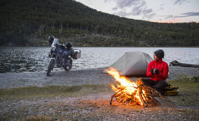 Frau genießt das Lagerfeuer im Camp neben einem stillen See in Feuerland - CAVF86140