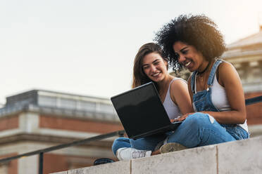 Weibliche Freunde verwenden Laptop, während sie auf einer Stützmauer sitzen - EYF08336