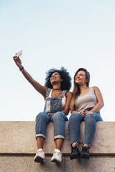 Weibliche Freunde, die ein Selfie machen, während sie auf einer Stützmauer gegen den klaren Himmel sitzen - EYF08339