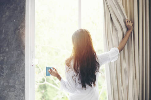 Junge Frau, die Kaffee trinkt, während sie den Vorhang vor dem Fenster zu Hause öffnet - EYF08400
