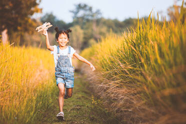 Porträt eines glücklichen Mädchens, das ein Modellflugzeug fliegt, während es auf einem grasbewachsenen Feld läuft - EYF08425
