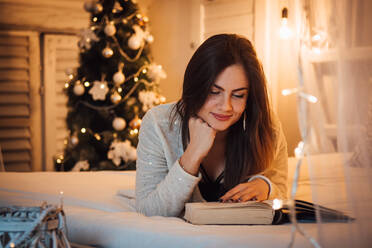 Junge Frau, die ein Buch liest, während sie auf dem Bett vor dem Weihnachtsbaum zu Hause liegt - EYF08449